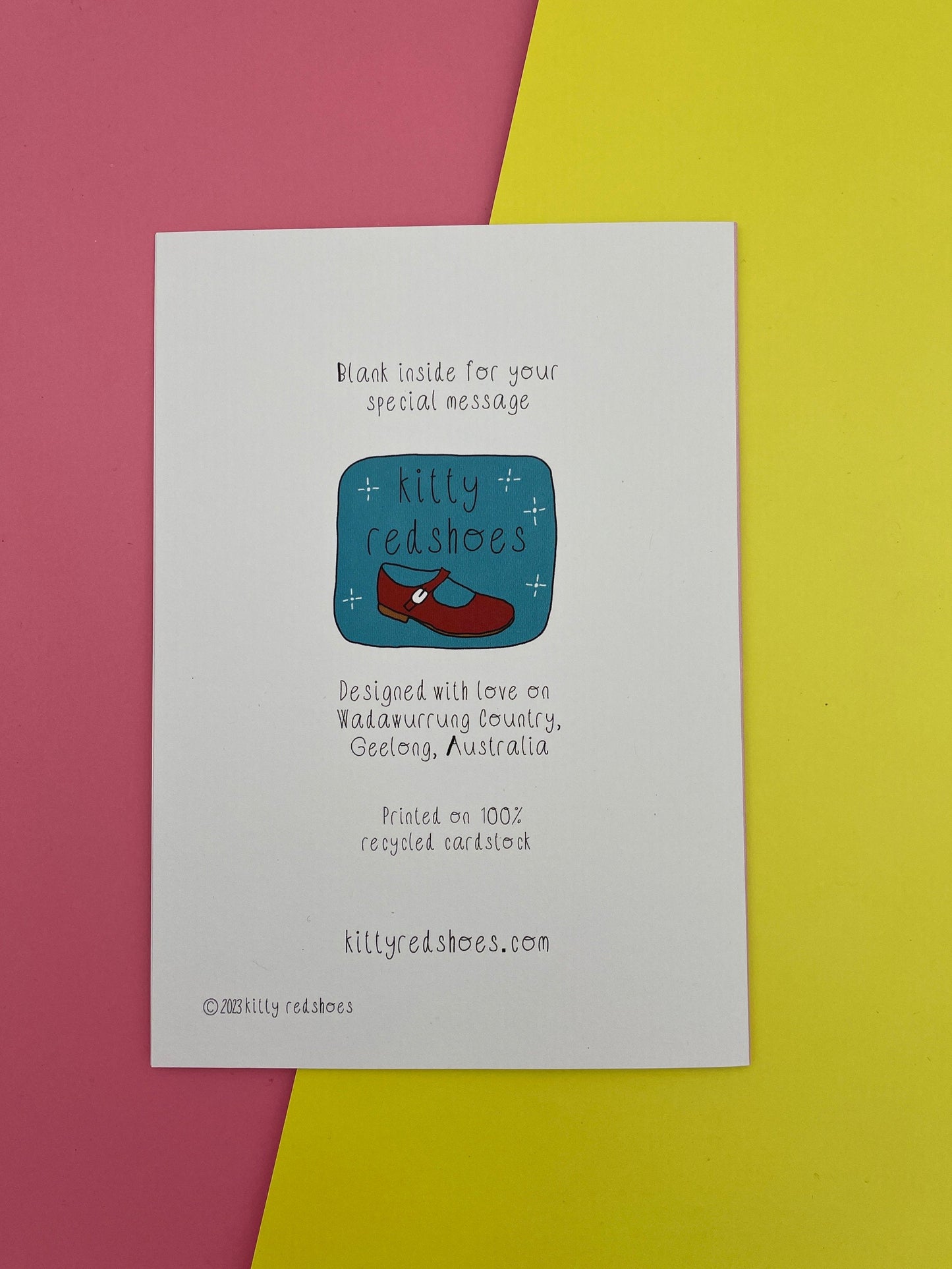 Reverse of greeting card with kitty redshoes logo and details about the card: Blank inside, Designed on Wadawurrung Country, Printed on 100% recycled cardstock, kittyredshoes.com, resting on a pink and yellow background.