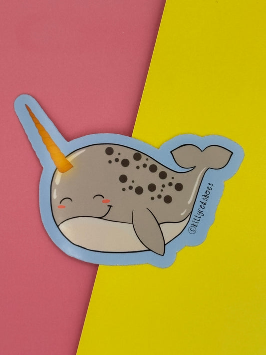 A sticker of a smiling grey cartoon narwhal with darker grey spots and a golden horn.