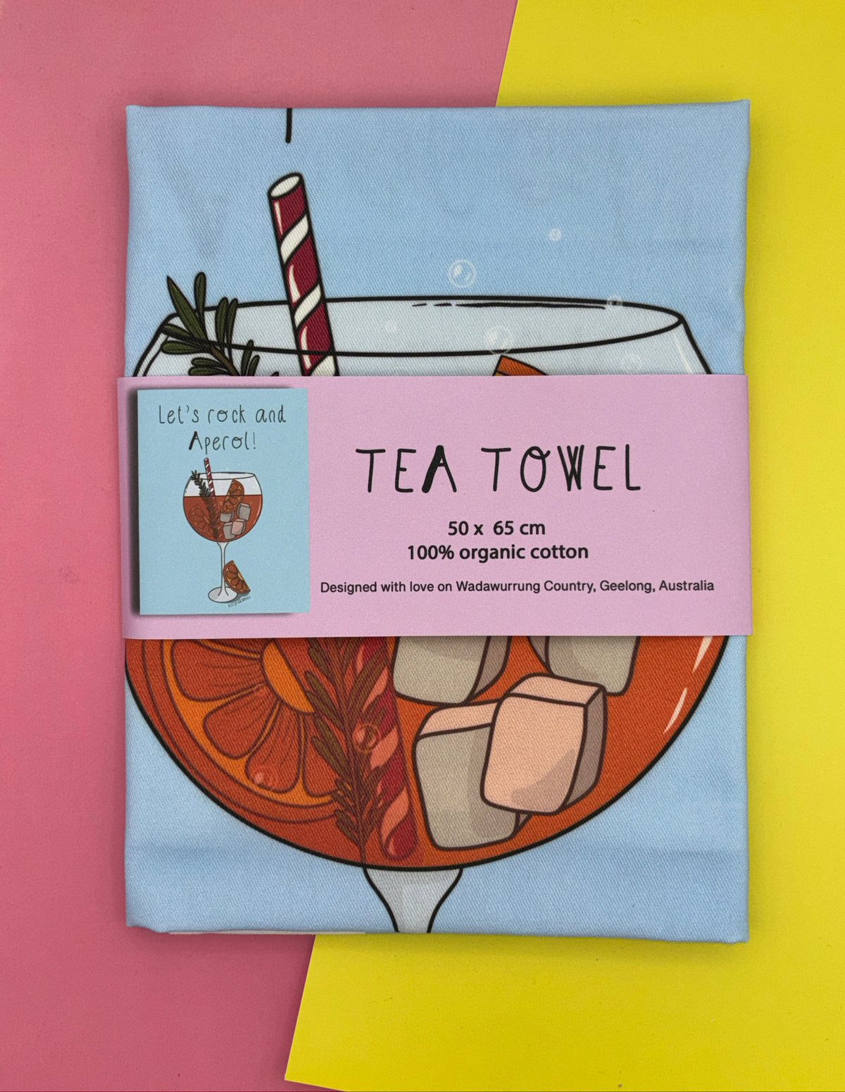 A folded tea towel with an illustration of an Aperol spritz glass on a yellow and pink background. The tea towel has a purple belly band around it with a picture of the 'Let's rock and Aperol' design, and text 50x65cm, 100%organic cotton and Designed with love on Wadawurrung Country, Geelong, Australia. 