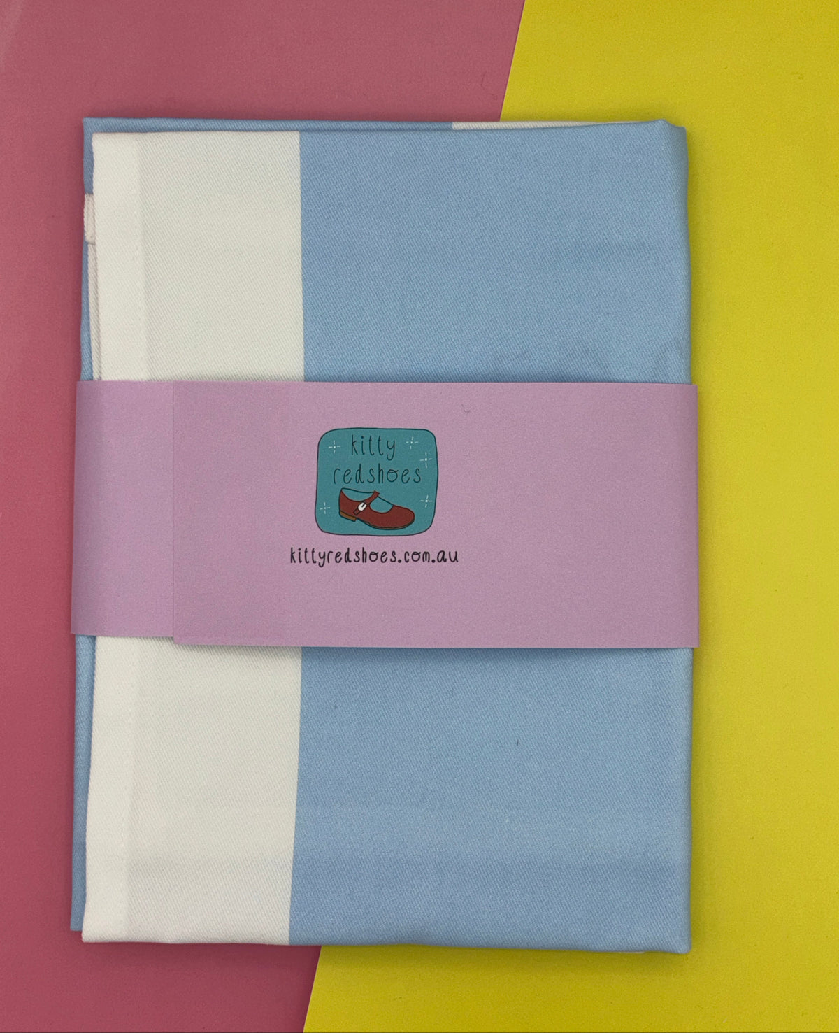 A folded blue and white tea towel on a pink and yellow background. The tea towel has a purple belly band with the kitty redshoes logo and website www.kittyredshoes.com.au printed on it.