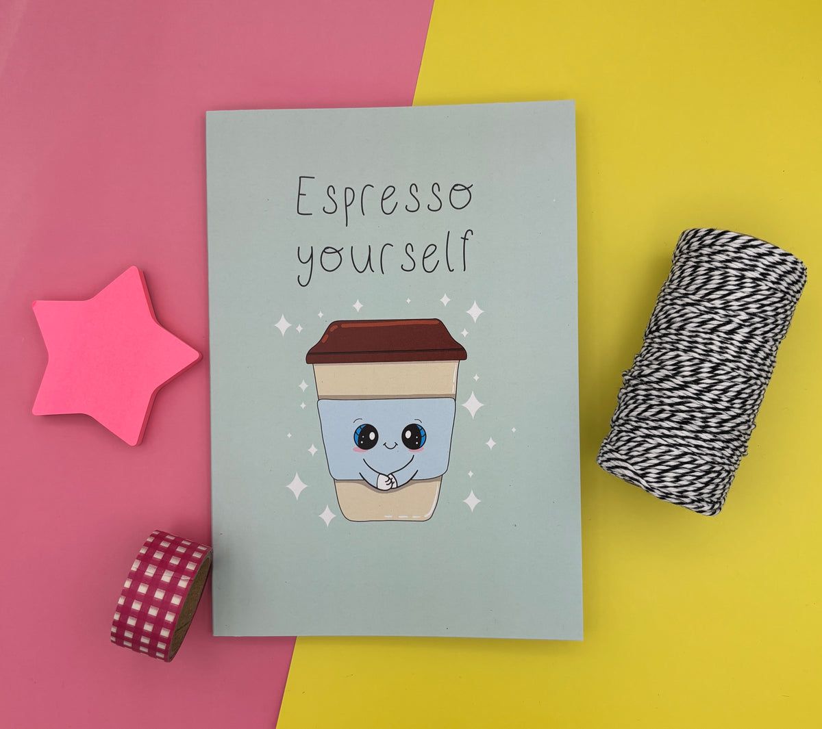A green notebook with the phrase 'Espresso yourself' above a cute cartoon coffee cup, styled on a split pink and yellow background with a pink star, washi tape, and string props.
