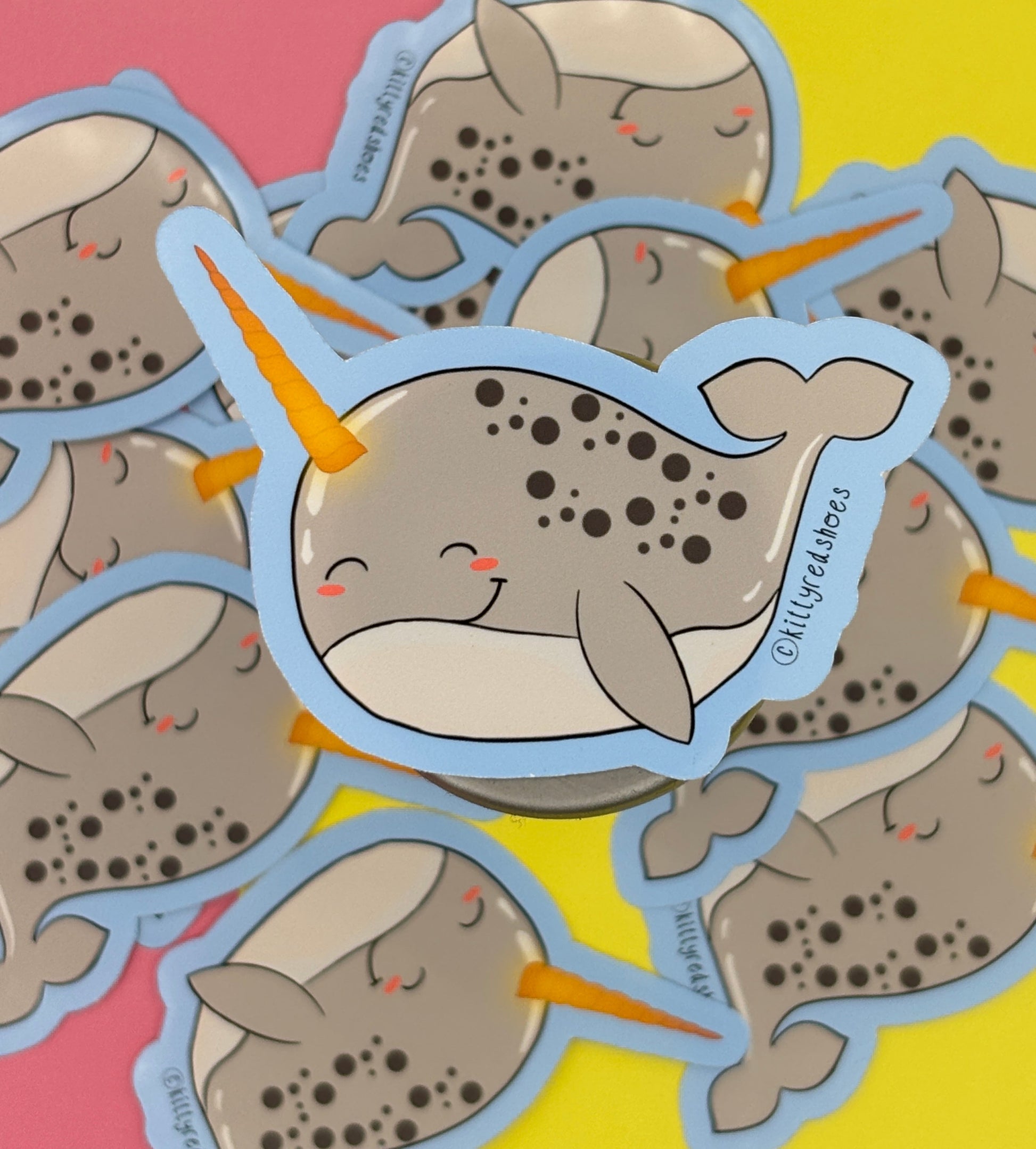 A sticker of a smiling grey cartoon narwhal with dark grey spots and a golden horn and a blue background.