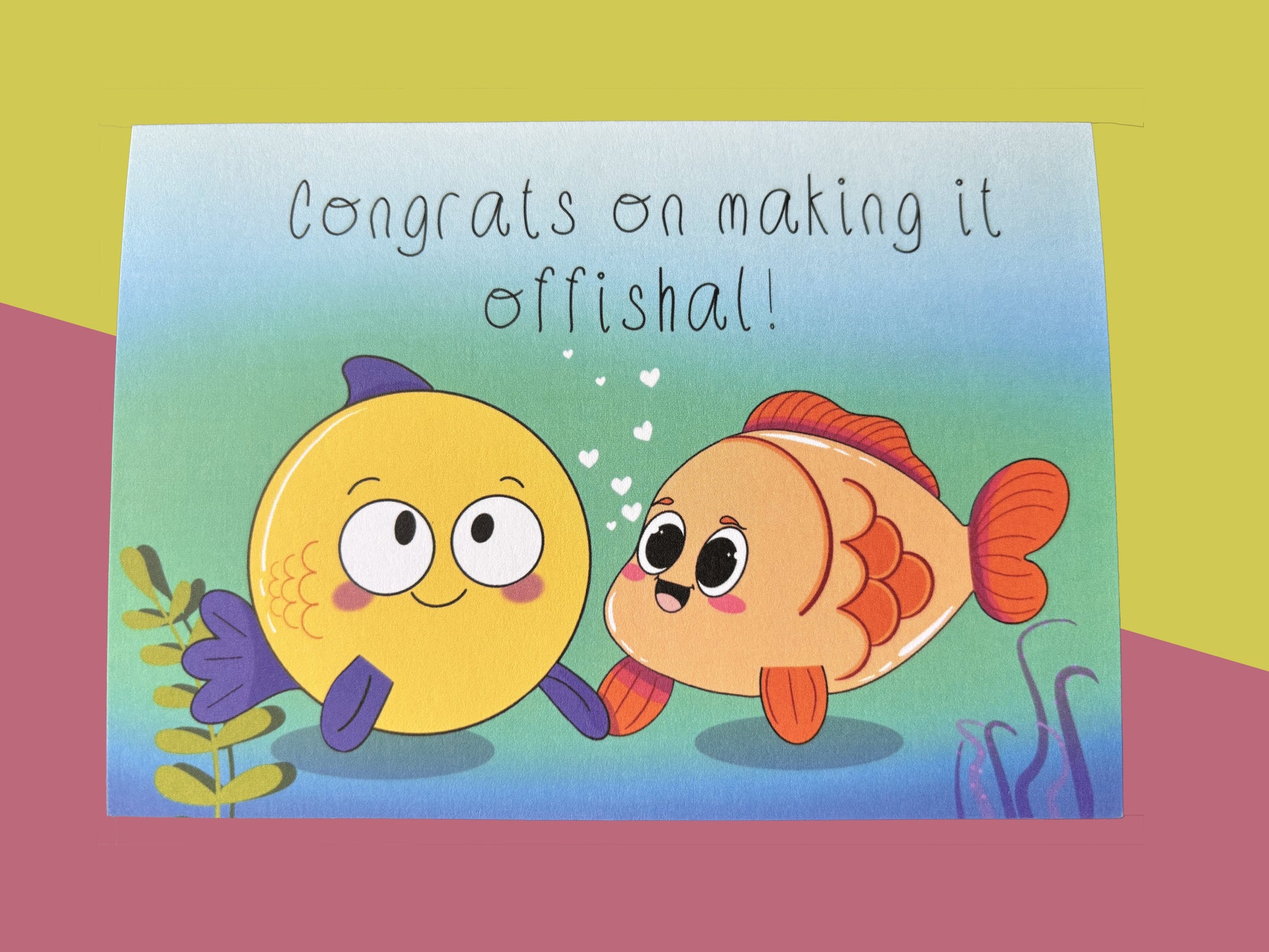 A colourful greeting card of 2 smiling cartoon fish holding fins under the sea, with white love hearts between them. Text: Congrats on making it offishal!