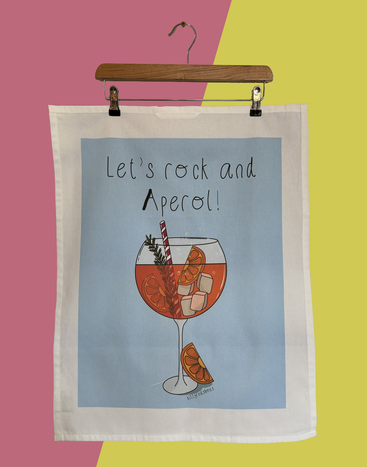 A blue tea towel with a printed illustration of a cocktail glass and the text 'Let's rock and Aperol!' hanging from a wooden hanger against a pink and yellow background.